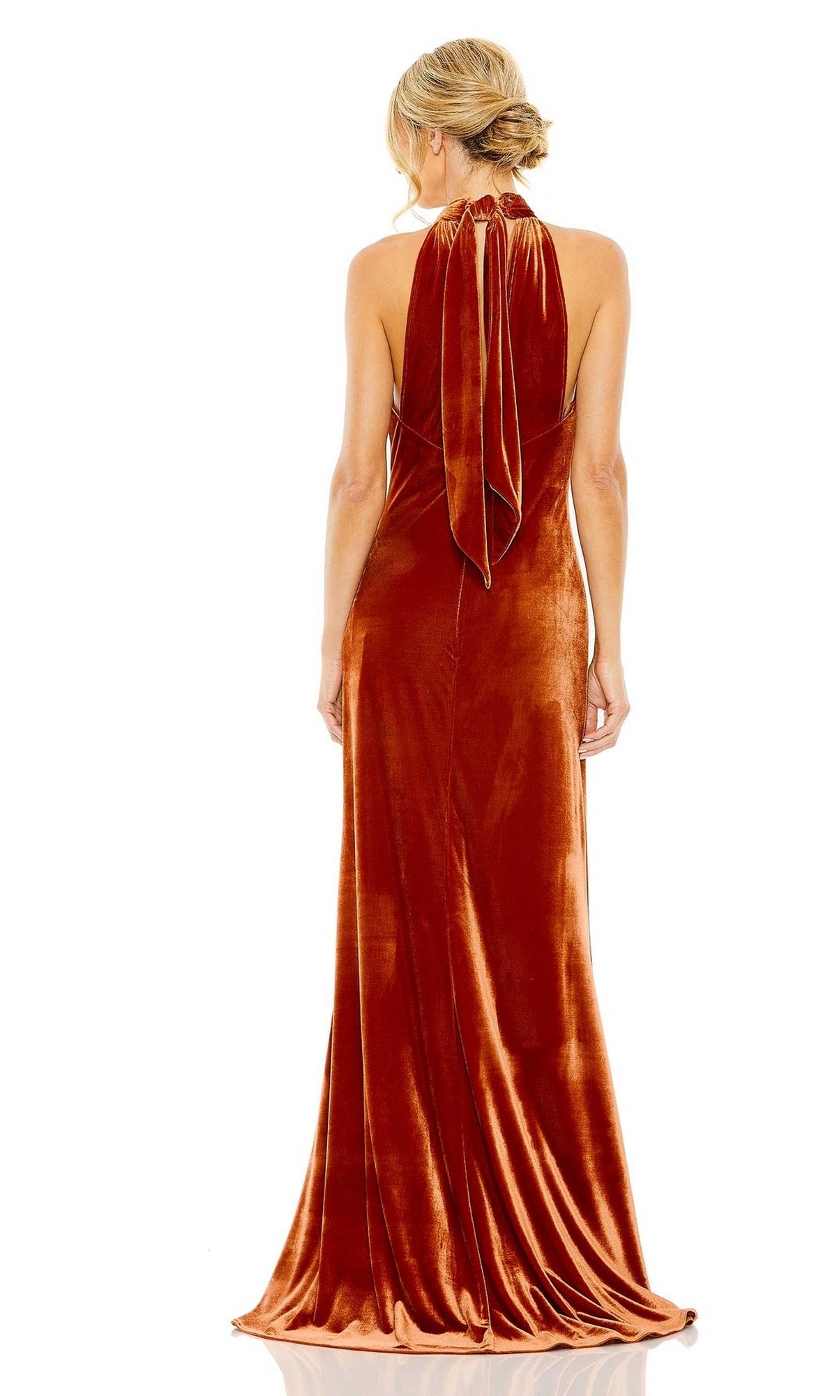 A woman with blonde hair in an updo models the Long Formal Dress 55954 by Mac Duggal—a floor-length burnt orange velvet gown with a high neck, open back, and draped tie detail. She is facing away from the camera.